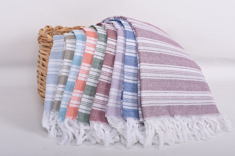 Puede incluir: Una colecci&oacute;n de toallas a rayas con flecos en varios colores, incluyendo azul, gris, naranja y morado, se exhiben junto a una cesta de mimbre. Las toallas tienen rayas horizontales blancas.