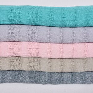 May include: Five woven cotton throws stacked on top of each other. The throws are in different colors: turquoise, light purple, pink, beige, and gray.
