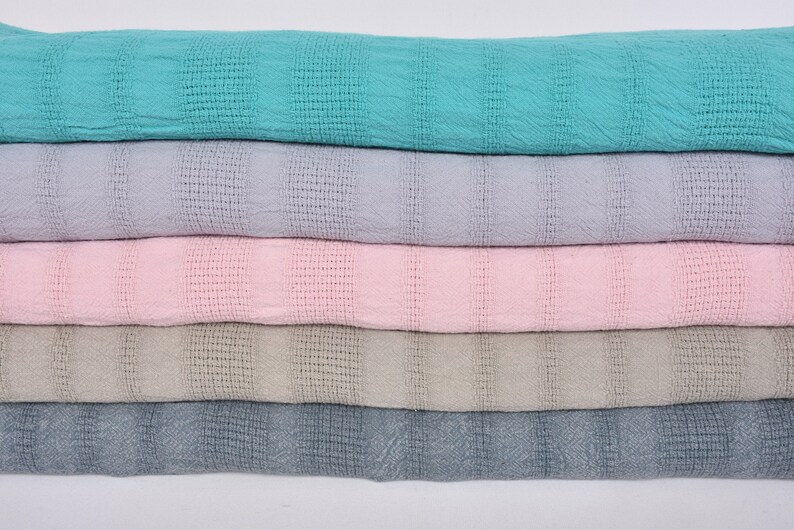 May include: Five woven cotton throws stacked on top of each other. The throws are in different colors: turquoise, light purple, pink, beige, and gray.