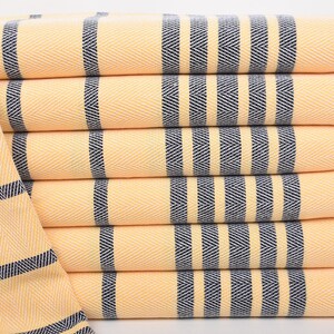 Può includere: Una pila di sei asciugamani a righe gialle e blu navy. Gli asciugamani hanno un motivo a spina di pesce e frange bianche alle estremità. Gli asciugamani sono ben piegati e impilati l'uno sull'altro.