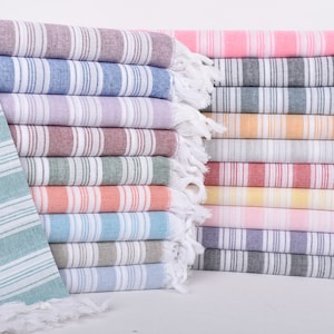 Puede incluir: Una pila de toallas turcas a rayas de colores. Las toallas son de varios colores, incluyendo rosa, azul, verde y naranja, con rayas blancas. Cada toalla tiene un borde con flecos.