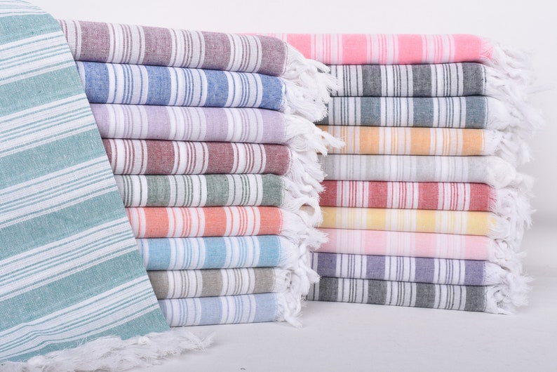 Puede incluir: Una pila de toallas turcas a rayas de colores. Las toallas son de varios colores, incluyendo rosa, azul, verde y naranja, con rayas blancas. Cada toalla tiene un borde con flecos.