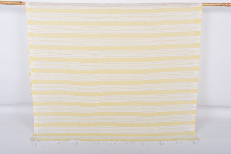 Puede incluir: Una toalla de playa a rayas amarillas y blancas colgada de una vara de madera. La toalla tiene rayas horizontales y un borde con flecos. La combinaci&oacute;n de colores es amarillo brillante y blanco.