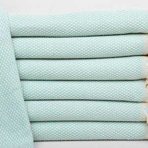 May include: A stack of light blue Turkish towels with white fringe. The towels are folded and stacked on a white background.