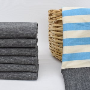 May include: A stack of six folded gray and white striped towels with a light blue stripe. A seventh towel with light blue and white stripes is in a woven basket. The towels are made of a soft, absorbent material.