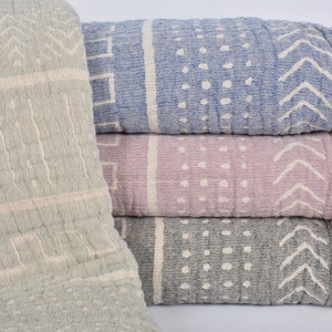 Puede incluir: Una pila de cuatro mantas de algodón con flecos. Las mantas presentan diferentes patrones geométricos en tonos de azul, gris y rosa.