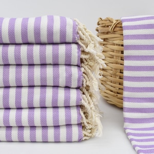 May include: A stack of six folded, striped towels in lavender and white. The towels have a fringe on the edge. A seventh towel is next to a woven basket. The towels are made of a soft, absorbent material.