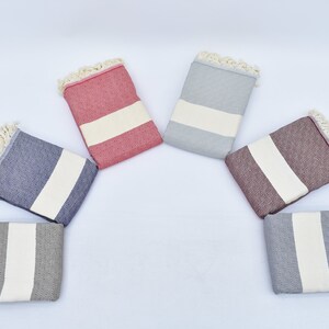 May include: Six Turkish towels in different colors, including gray, blue, red, and brown. Each towel has a white stripe in the center and fringe around the edges.