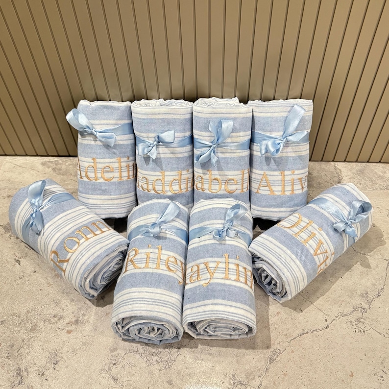 Puede incluir: Toallas enrolladas a rayas azules y blancas, atadas con cintas azul claro. Cada toalla est&aacute; bordada con un nombre en hilo dorado. La disposici&oacute;n sugiere un regalo o art&iacute;culo personalizado, presentado sobre una superficie de m&aacute;rmol.