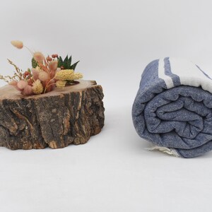 Op de afbeelding: Een opgerolde blauwe en witte gestreepte strandhanddoek met witte strepen. De handdoek is strak opgerold en ligt op een wit oppervlak.