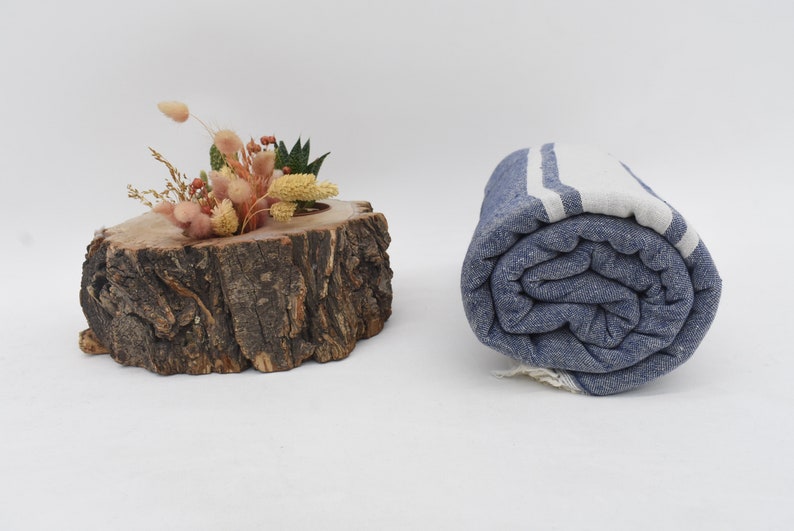 Op de afbeelding: Een opgerolde blauwe en witte gestreepte strandhanddoek met witte strepen. De handdoek is strak opgerold en ligt op een wit oppervlak.
