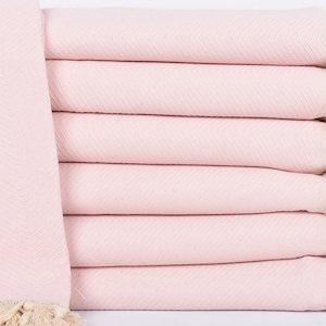 May include: A stack of six pale pink herringbone-patterned towels with cream-coloured fringe. The towels are neatly folded, showing the texture and colour. The fringe adds a decorative element to the towels.