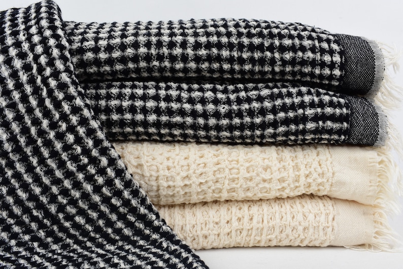 May include: A stack of four waffle weave towels in black and white and natural colors. The towels are folded and have a textured waffle weave pattern.