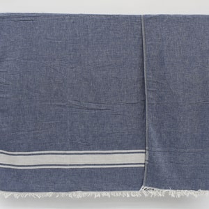 Op de afbeelding: Een blauwe en witte gestreepte katoenen deken met franjes aan de randen. De deken hangt aan een houten stok.