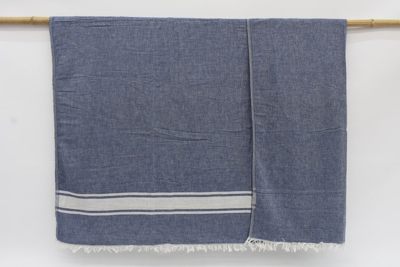 Op de afbeelding: Een blauwe en witte gestreepte katoenen deken met franjes aan de randen. De deken hangt aan een houten stok.