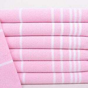 Può includere: Una pila di sei asciugamani rosa e bianchi a righe. Gli asciugamani sono piegati con cura, con frange bianche visibili sui bordi. Gli asciugamani sono realizzati in un materiale morbido e tessuto.
