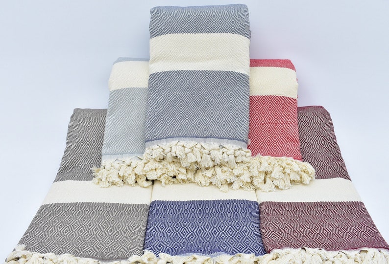 May include: A set of six Turkish towels in various colors, including gray, blue, and red. The towels are made of cotton and have a fringed edge. They are stacked on top of each other, with the top towel folded in half.