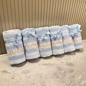 Puede incluir: Mantas enrolladas a rayas azules y blancas atadas con cintas azul claro. Cada manta tiene un nombre bordado en color dorado. Las mantas est&aacute;n dispuestas en fila, sugiriendo un regalo o un art&iacute;culo personalizado.