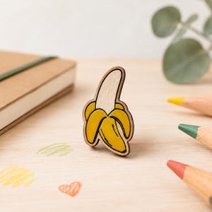Puede incluir: Un broche de madera con forma de plátano parcialmente pelado. El plátano presenta una cáscara de color amarillo brillante y una fruta interior de color beige claro, delineada con líneas de color marrón oscuro. El diseño es plano y liso.