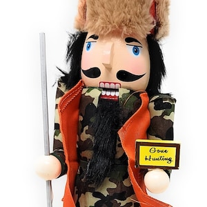 May include: A wooden nutcracker in a camouflage outfit with an orange vest and a furry hat. The nutcracker holds a silver pole and a sign that says "Gone Hunting". The base of the nutcracker says "DAD".