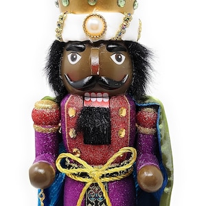 May include: A wooden nutcracker with a purple and red robe, a gold crown, and a gold beard. The nutcracker is holding a gold orb and is standing on a purple base with the word "STEPHANIE" written on it.