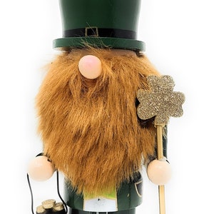 May include: A wooden figurine of a leprechaun wearing a green hat and holding a pot of gold and a shamrock. The figurine is standing on a green base with the word "JOSI" written on it.