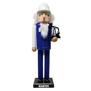 May include: A wooden nutcracker in the form of a construction worker, wearing a white hard hat, blue uniform, and holding a black oil derrick. The nutcracker has a white beard and mustache, and the base reads "MARTIN".
