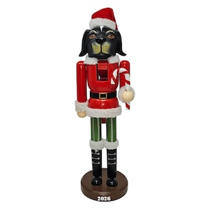 May include: A Christmas-themed dog nutcracker figurine. The dog has a black face and ears, wearing a red Santa hat and suit with white trim. It holds a candy cane and stands on a brown base with the number "2026".