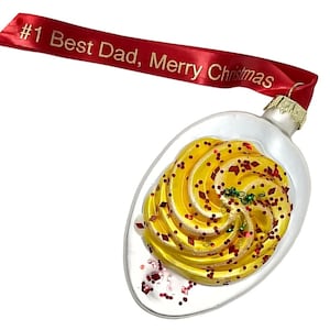 May include: A clear glass Christmas ornament shaped like a spoon, filled with yellow swirls and red glitter. A red ribbon with the text "#1 Best Dad, Merry Christmas" is attached to the ornament.