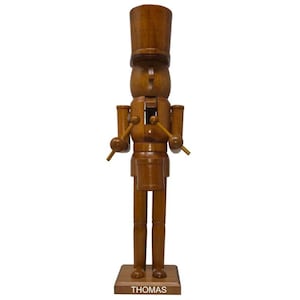 May include: A tall, brown wooden nutcracker figure wearing a cylindrical hat. The figure holds two drumsticks and stands behind a small drum. The base of the nutcracker is rectangular and features the name THOMAS printed in white capital letters.