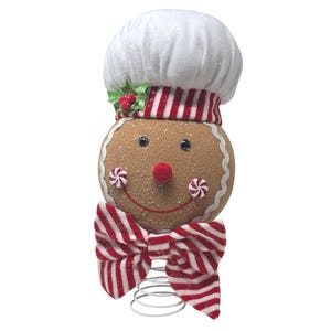 May include: A gingerbread person tree topper featuring a white chef hat with red and white striped trim, a round tan face with black eyes, a red pom-pom nose, peppermint candy cheeks, a red stitched smile, and a red and white striped bow tie.