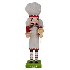 May include: A wooden chef nutcracker figurine. The figurine wears a white chef's hat with red trim, a red oven mitt, and a white apron. It holds a wooden plate and a spoon. The base is green with the name "PHILLIP" in white.