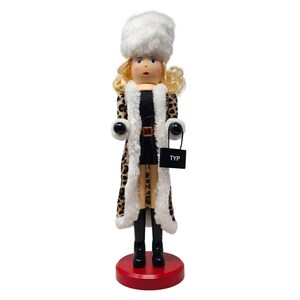 May include: A wooden nutcracker doll with blonde curly hair, blue eyes, and red lips. The doll wears a white fur hat and a leopard print coat with white trim. It holds a black purse with the letters "TYP" on it and stands on a red base.