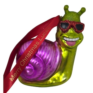 May include: A green snail ornament with a shiny purple shell, wearing red glittery sunglasses and sticking out a pink tongue. A red ribbon is attached to the top, featuring the gold printed text Merry Christmas Lucas.