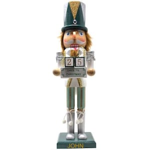 May include: A nutcracker figurine wearing a green and silver hat with gold accents, a white jacket with gold trim, and silver boots. It holds a sign with two blocks displaying the number 25 and the text DAYS TIL CHRISTMAS. The base is labeled JOHN.