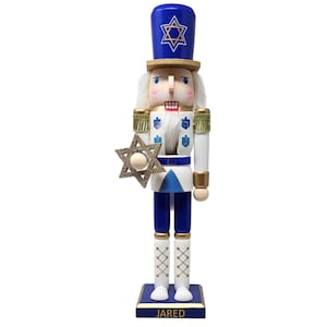 May include: A wooden nutcracker with a white beard and blue and gold uniform. The nutcracker is holding a gold Star of David and has a blue hat with a Star of David on it. The base of the nutcracker is blue and has the name "JARED" written on it.