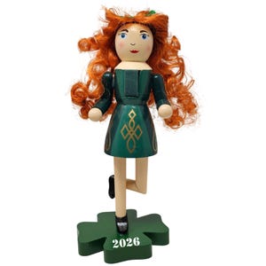 May include: Wooden doll with red curly hair, wearing a green dress with gold accents. The doll is standing on a green shamrock base with the number 2026. The doll has a painted face with blue eyes and a red mouth.