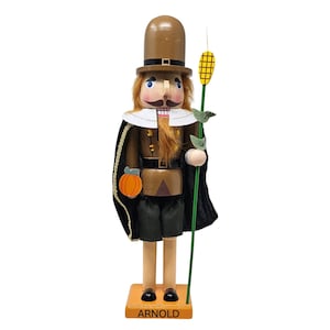 May include: A wooden nutcracker figurine in the shape of a pilgrim, wearing a brown top hat, coat, and shorts. The figurine holds a corn stalk and a pumpkin. The base of the figurine is orange and reads "ARNOLD".
