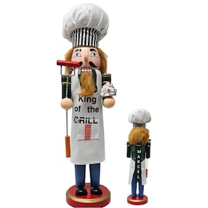 May include: Two wooden nutcrackers in the shape of chefs. The larger one holds a grill tool and a plate of food, and wears an apron that says "King of the Grill". The smaller one has the name "Marcel" on the back.