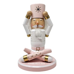 May include: A pink and white wooden nutcracker figurine with a smiling face, white beard, and a pink hat with a black star design. The figurine has gold accents and is standing on a pink base with the text "ZOMBA HILLS 2025".