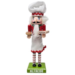 May include: A wooden nutcracker in the shape of a chef, holding a pizza and a pizza box. The chef wears a white hat, apron, and has a white beard. The base is green with the name "ALFREDO" in white.