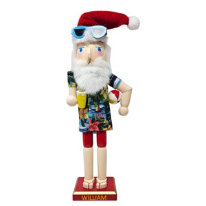 May include: A wooden nutcracker of Santa Claus wearing a red hat, sunglasses, a tropical print shirt, red shorts, and holding a beach ball and a yellow drink. The base of the nutcracker is red and has the name "WILLIAM" written on it.