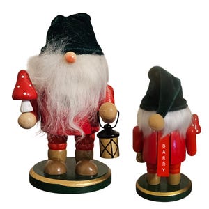 May include: Two wooden gnome figurines. The larger gnome has a red coat, white beard, and a green velvet hat. It holds a mushroom and a lantern. The smaller gnome has a red coat, white beard, and a green velvet hat. The name "Barry" is printed on a tag.