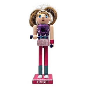 May include: A wooden nutcracker doll in the shape of a woman, with brown hair in pigtails, wearing a purple top with white triangle accents, a pink glitter skirt, and pink leggings. The doll is holding blue dumbbells and has the name "Kimber" on the base.