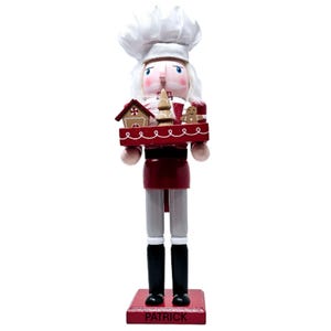 May include: A wooden nutcracker in the form of a chef, wearing a white hat and a red apron. The nutcracker holds a tray with gingerbread house, tree, and man decorations. The base is red with the name "PATRICK".