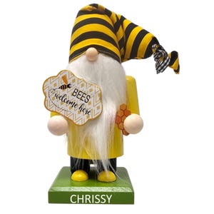 May include: A yellow wooden gnome with a white beard and a black and yellow striped hat. The gnome is holding a sign that says "BEES welcome here" and a honeycomb. The gnome is standing on a green base with the name "CHRISSY" written on it.