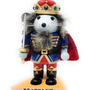 May include: A decorative mouse nutcracker wearing a blue and red uniform with gold accents, a red cape, and a crown. The mouse holds a sword and stands on a cheese-shaped base with the name "FRANKLYY".