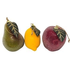 May include: Three fruit-shaped ornaments featuring a glittery green pear, a bright yellow lemon, and a deep red apple. Each piece includes a dark green leaf accent with gold vein detailing and a small brown string loop for hanging purposes.
