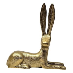 May include: A gold-colored, decorative hare figurine. The hare is in a reclining position with its long ears upright. The sculpture has a textured surface and is set against a white background.