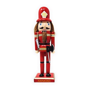 May include: A wooden firefighter nutcracker with a red uniform, a black axe, and a yellow "FD" on his hat. The base of the nutcracker is painted gold with the name "RAYMOND" in red letters.